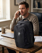 Man on a phone call at a home office desk with a black Grit With Faith embroidered tech backpack